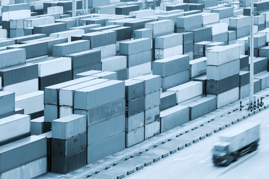 Rows of stacked shipping containers in an industrial yard, with a blurred truck driving along the road in the foreground.